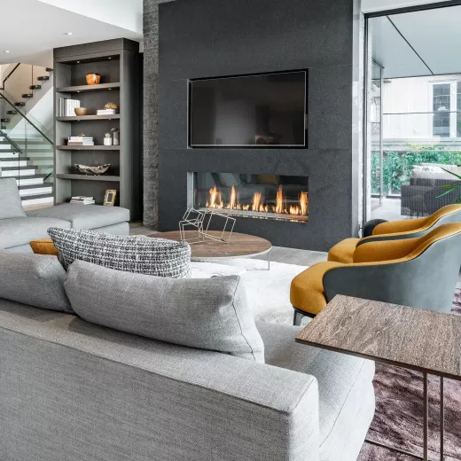Modern living room with a large fireplace, gray sofas, patterned armchairs, and built-in shelves. Glass walls reveal a stairway and outdoor view.