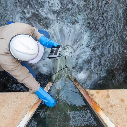 A worker in protective gear uses a device to monitor water quality in a flowing channel, with water splashing as it interacts with the sensor.