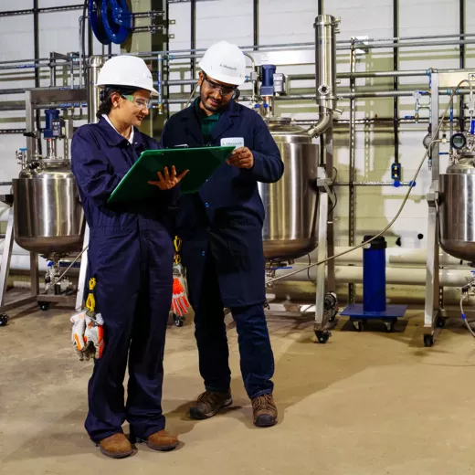Two engineers in safety gear, one male and one female, are discussing a document in an industrial facility with steel tanks and piping in the background.