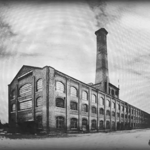 A black and white photo of an old industrial building with a tall chimney, featuring large, arched windows under a dramatic cloudy sky.