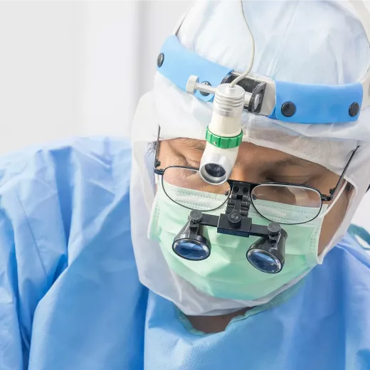 A focused healthcare professional wearing surgical scrubs, a headlamp, magnifying loupes, and a surgical mask, preparing for a medical procedure.
