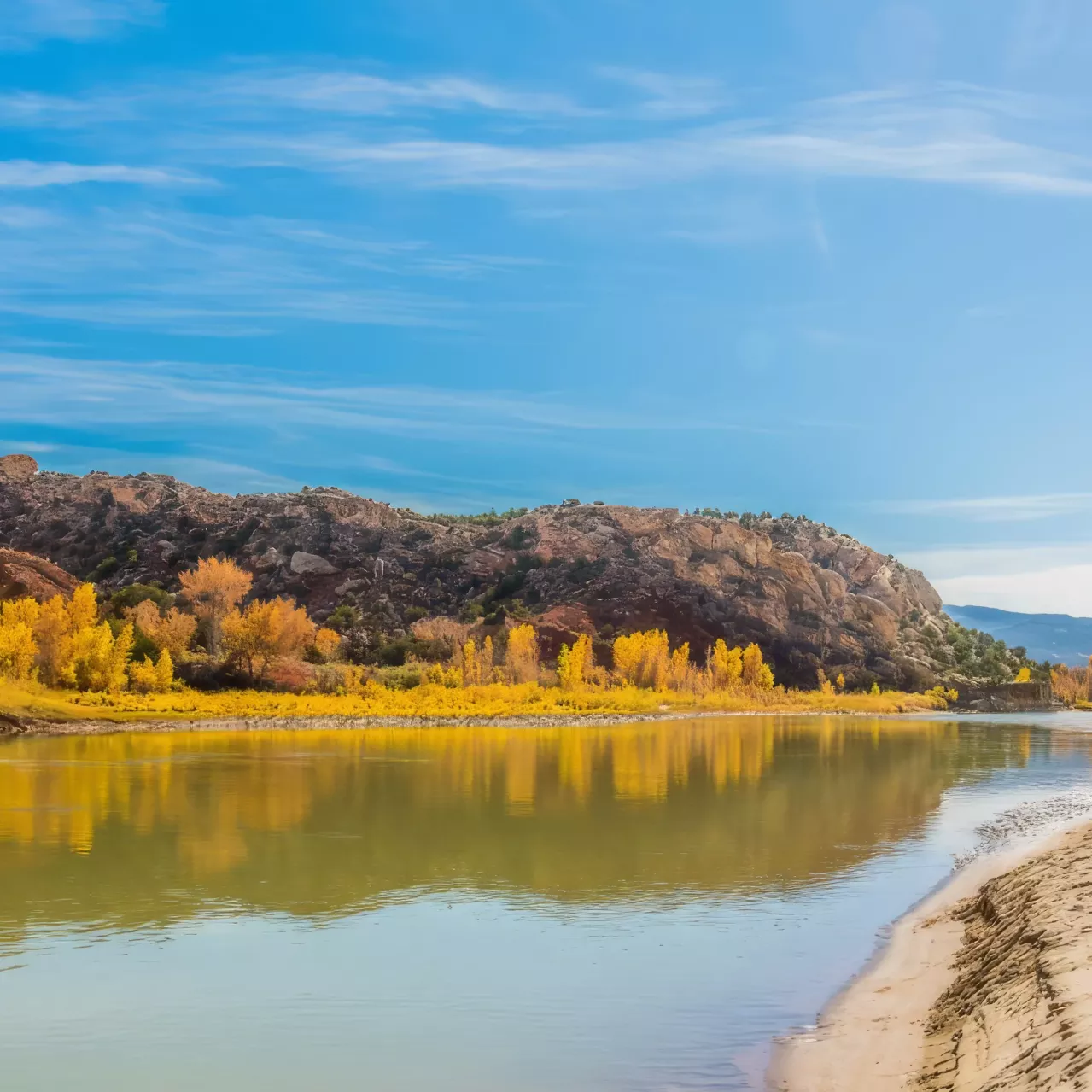 Tranquil river scene with a sandy shore in the foreground, reflective water, and a rocky hill with autumn-colored trees under a clear sky.