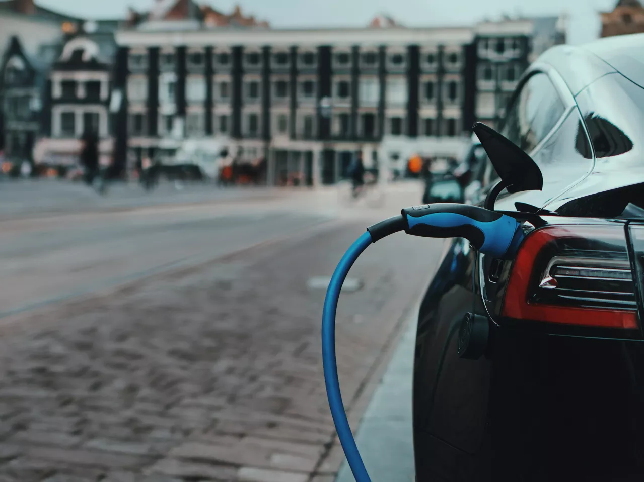 An electric car being charged on a city street, with the charging cable connected to the car's port and blurred buildings in the background.
