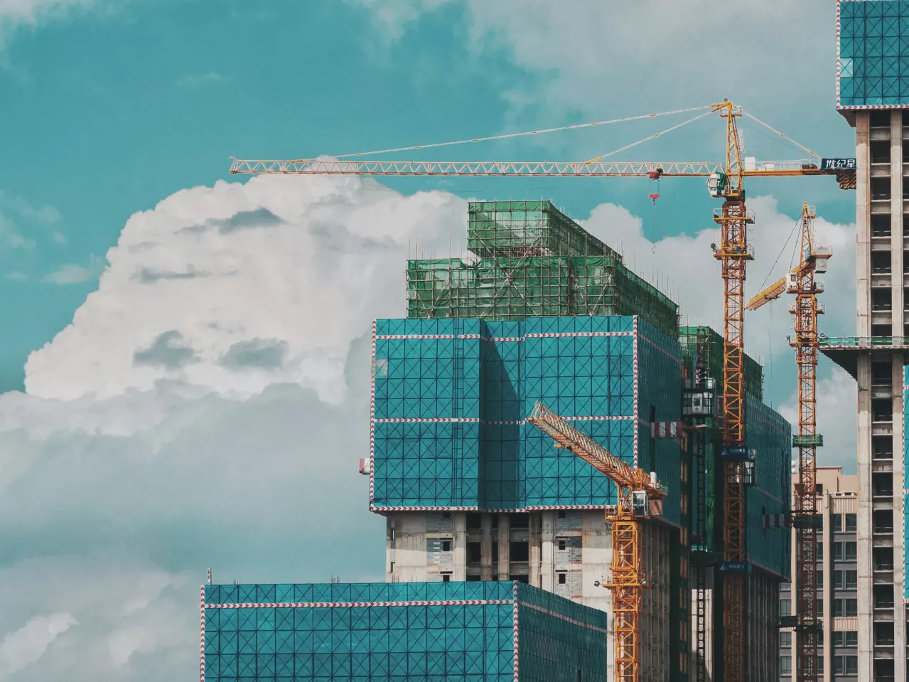 High-rise buildings under construction with scaffolding, covered in green mesh, and large yellow cranes against a cloudy sky.