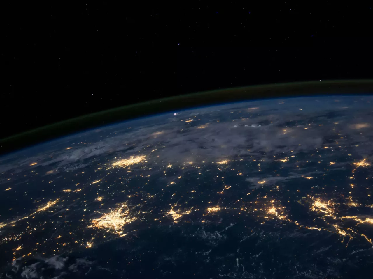 A view of earth from space at night showing the planet's curved horizon. Some clouds are illuminated by city lights. Clusters of city lights showing in certain areas looking like connected light streams.