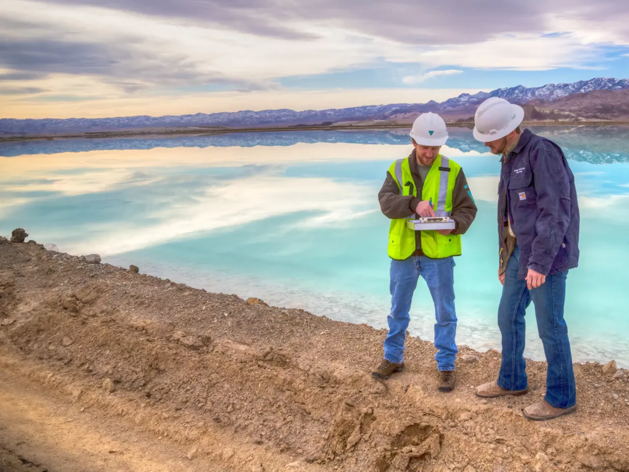 Two engineers in hard hats examine documents at a salt flat with mountains in the background, indicating a renewable energy project focusing on lithium extraction.