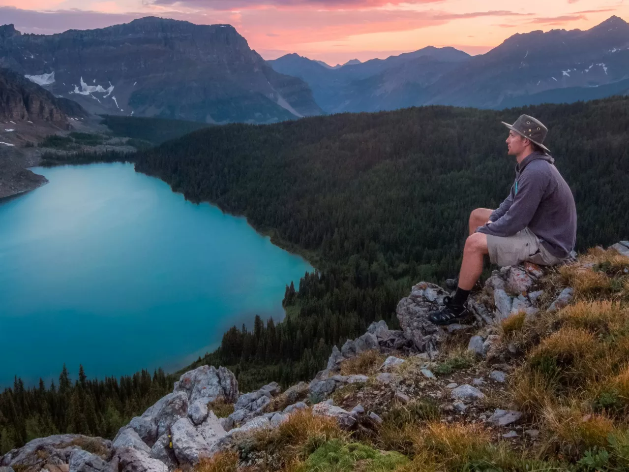 A person wearing a hat, gray hoodie, and shorts sits on a rocky ledge overlooking a blue lake surrounded by dense forest and mountains. The sun is setting in the background, casting a warm glow across the sky.