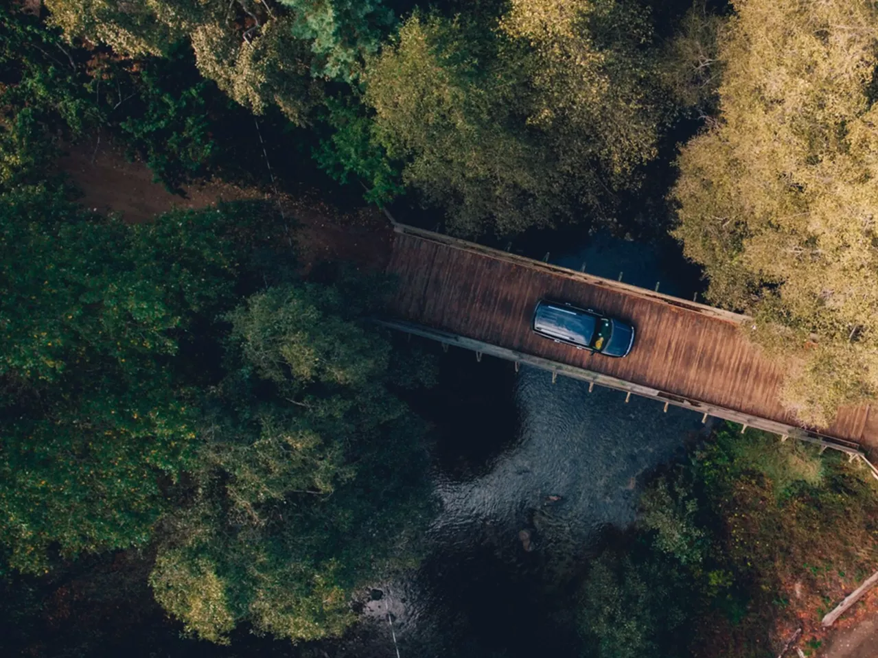 Aerial view of a blue car crossing a narrow wooden bridge over a river, surrounded by dense green trees.