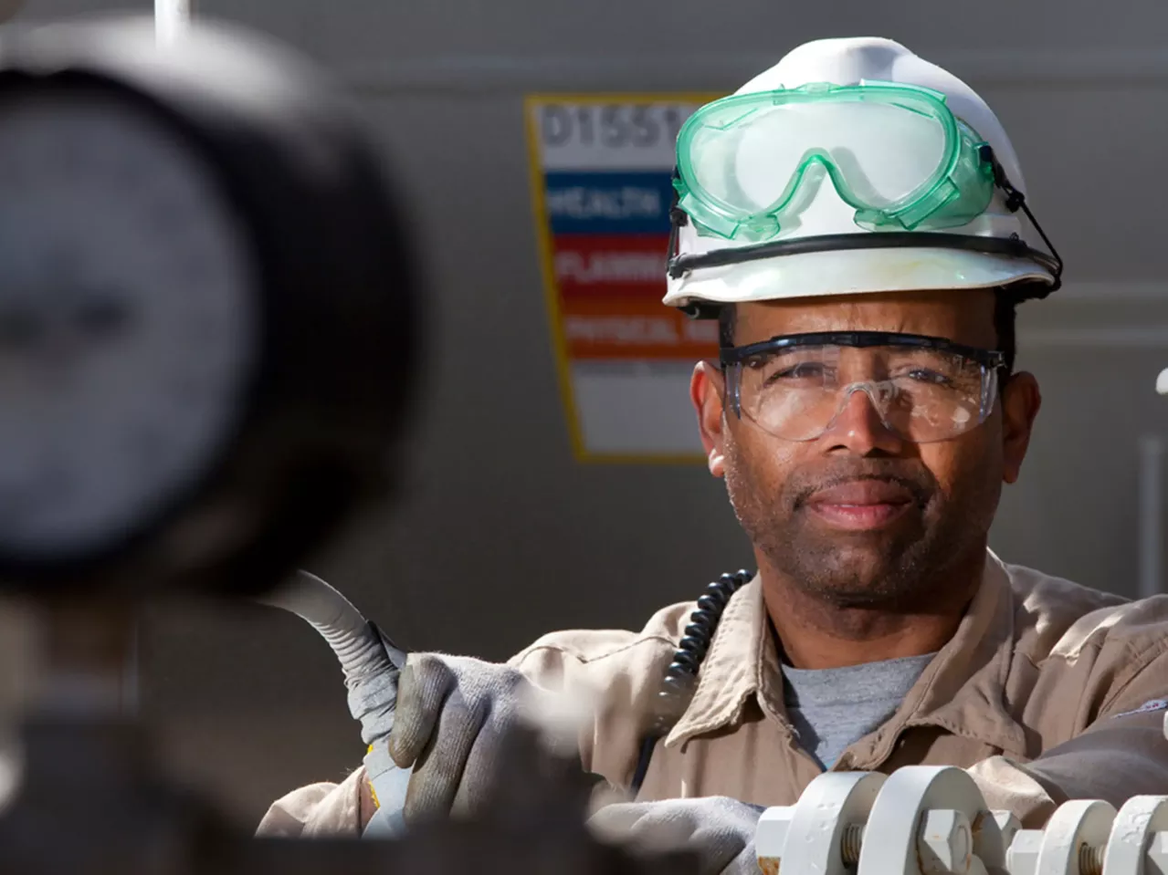 A worker wearing a white hard hat, safety goggles, and gloves operates equipment in an industrial setting. He stands behind machinery, maintaining a focused expression, with other industrial components and a gauge partially visible in the foreground.