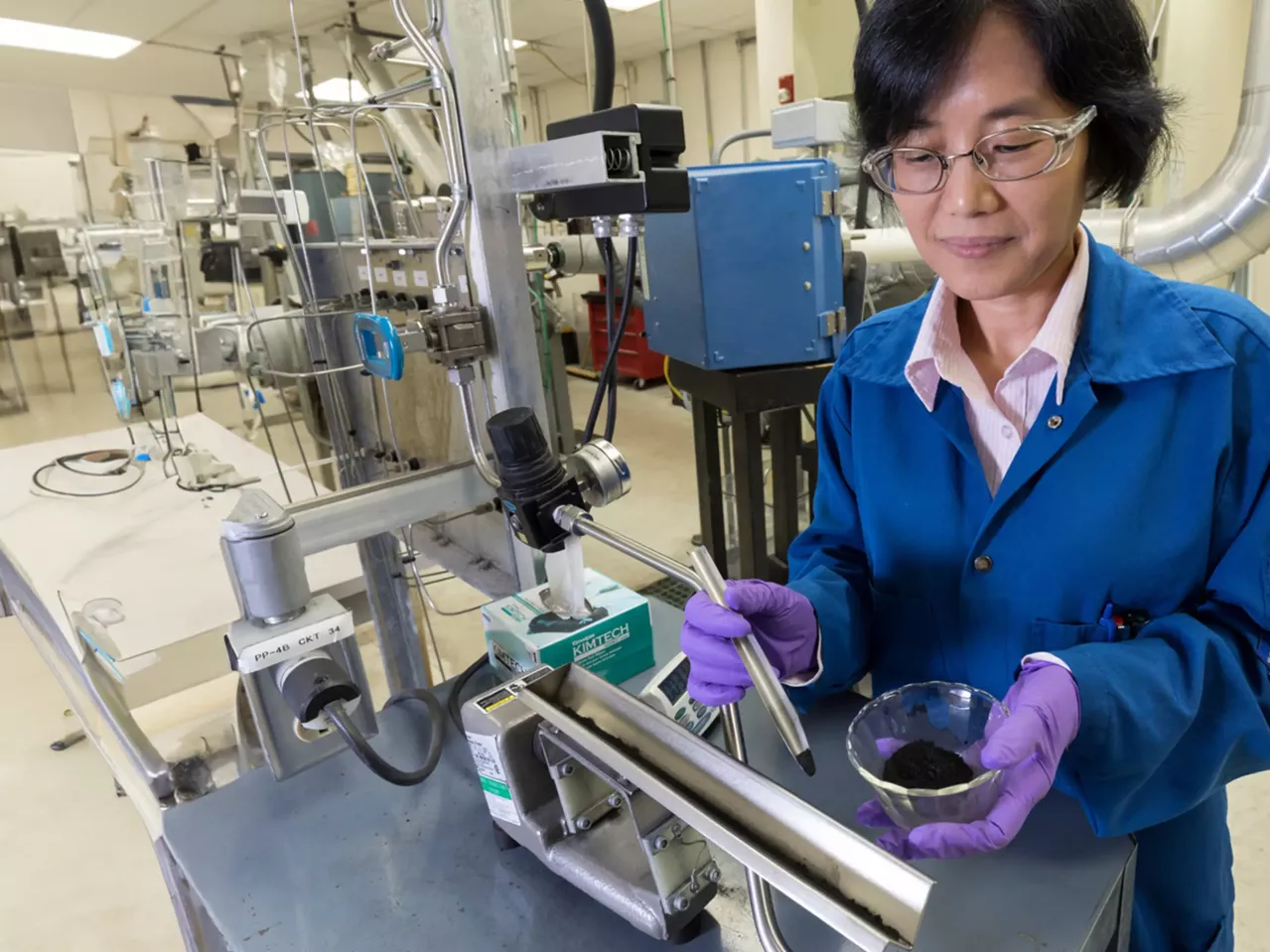 A female scientist in a lab coat conducts an experiment in an industrial setting, interacting with equipment while examining a sample in a mixing dish.